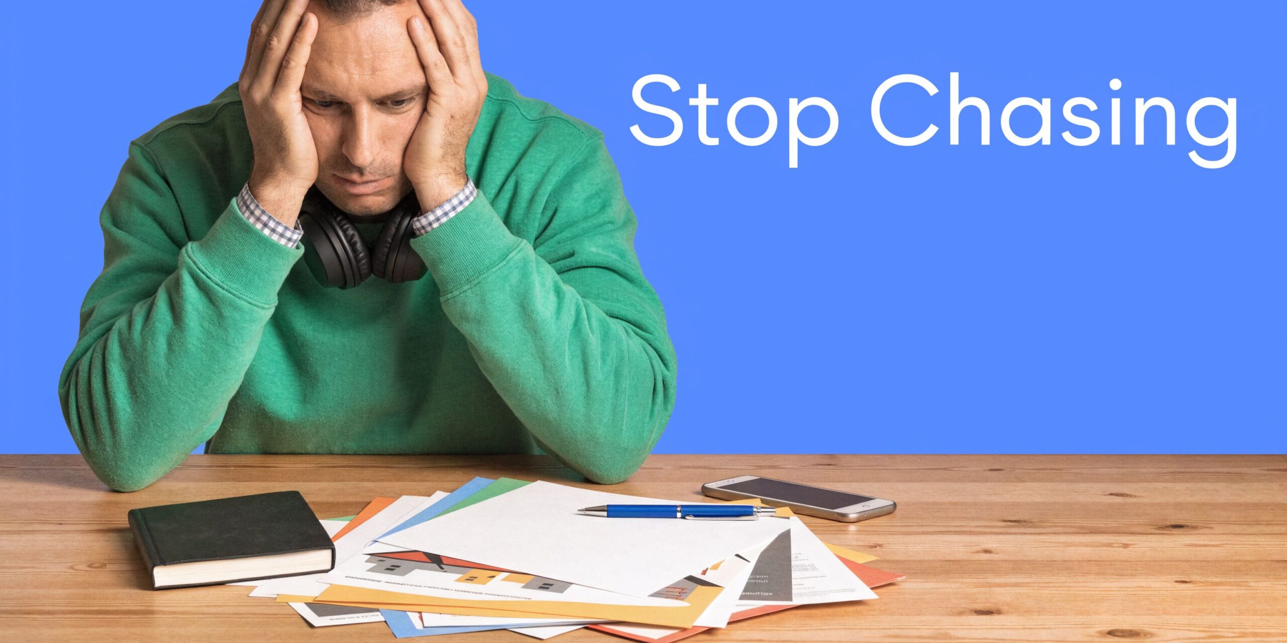 Stressed man sitting at a wooden desk with construction documents and a phone on a blue background.