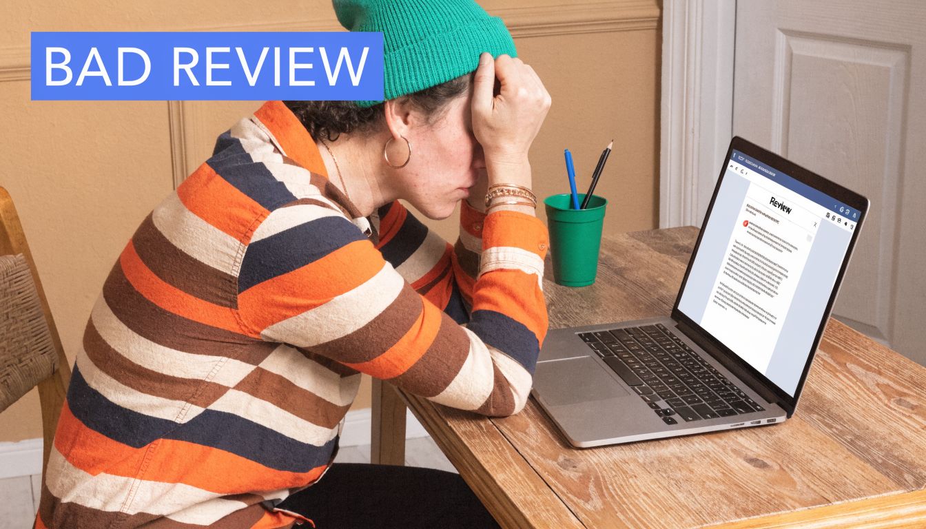 A person sitting at a desk looking stressed while reading a negative review on their laptop.